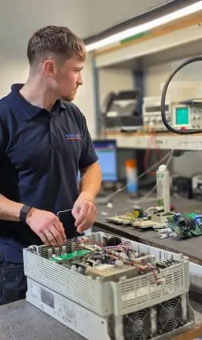 Engineer at Neutronic Technologies working on an electronic device with exposed components at a workstation equipped with tools and a computer monitor.