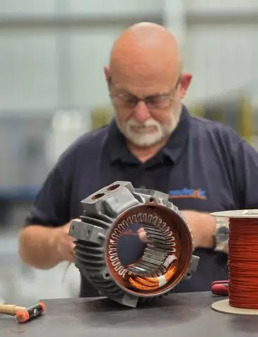 Person in a dark shirt working on an electric motor stator with visible copper windings, alongside a spool of copper wire and tools like a hammer on the workbench.