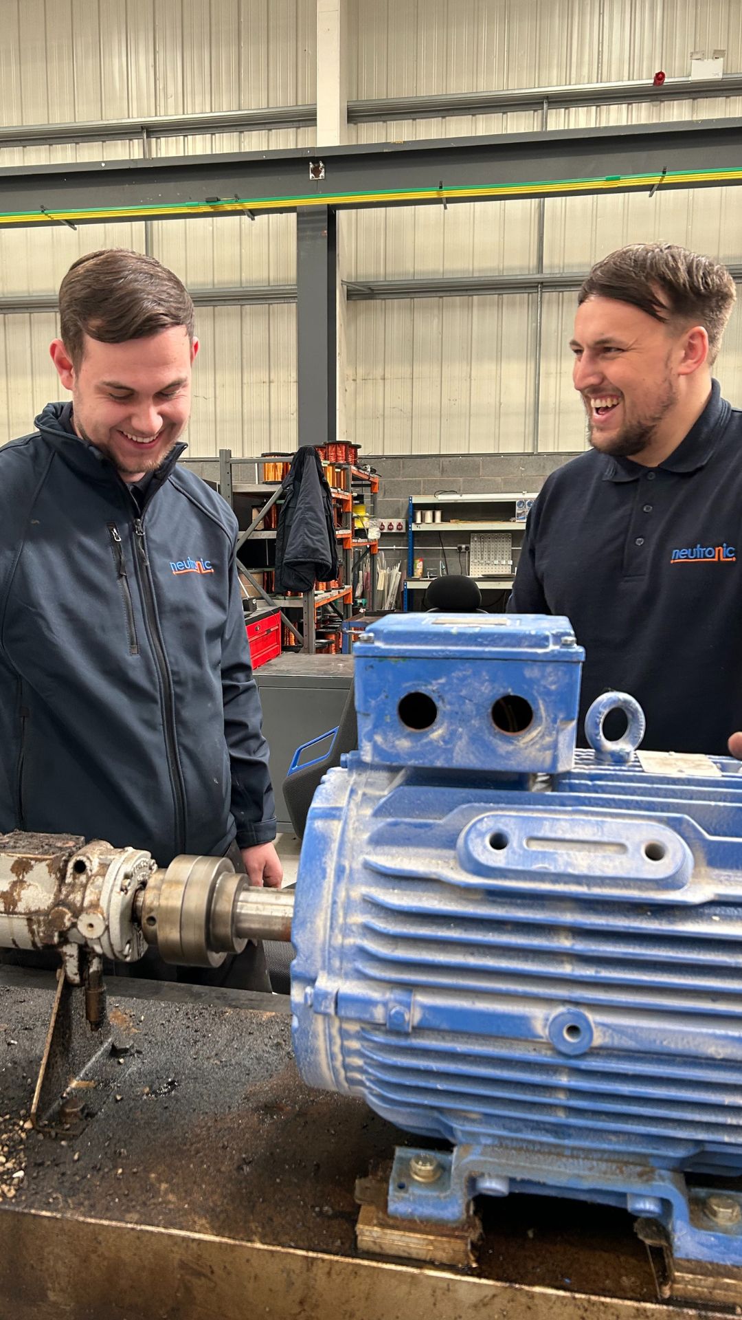 Two individuals in a workshop repairing a large blue industrial pump and motor assembly on a workbench, with tools and equipment visible in the background.