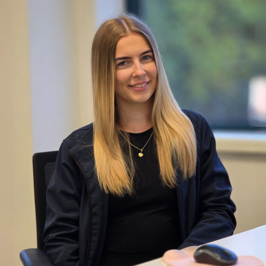 Ellie Gamble, Operations Co-ordinator at Neutronic Technologies, seated on an office chair, wearing a black top and dark jacket with a small pendant necklace. Her face is intentionally blurred.