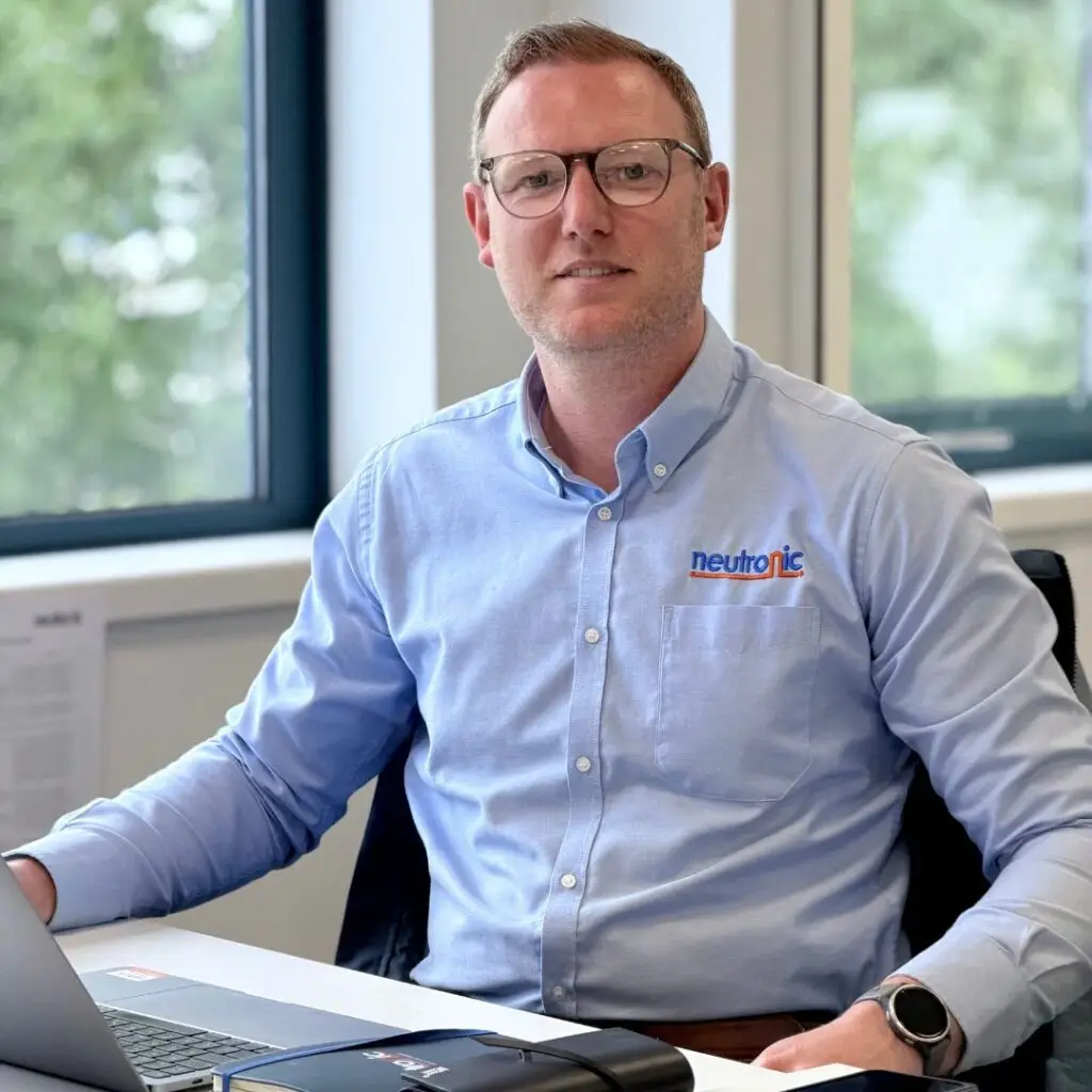 Neil Gallant, managing director of Neutronic Technologies Ltd, at his office desk in a light blue company shirt. He’s working on a computer, has a watch on his left wrist. The office has big windows with green things outside.